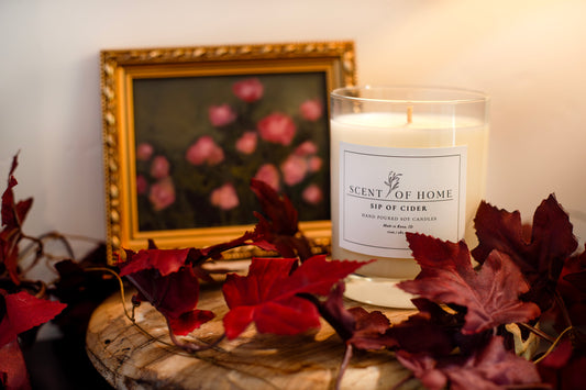 Scent of Home Sip of Cider candle surrounded by autumn leaves and a vintage floral painting, styled to reflect the brand’s cozy and artistic inspiration.