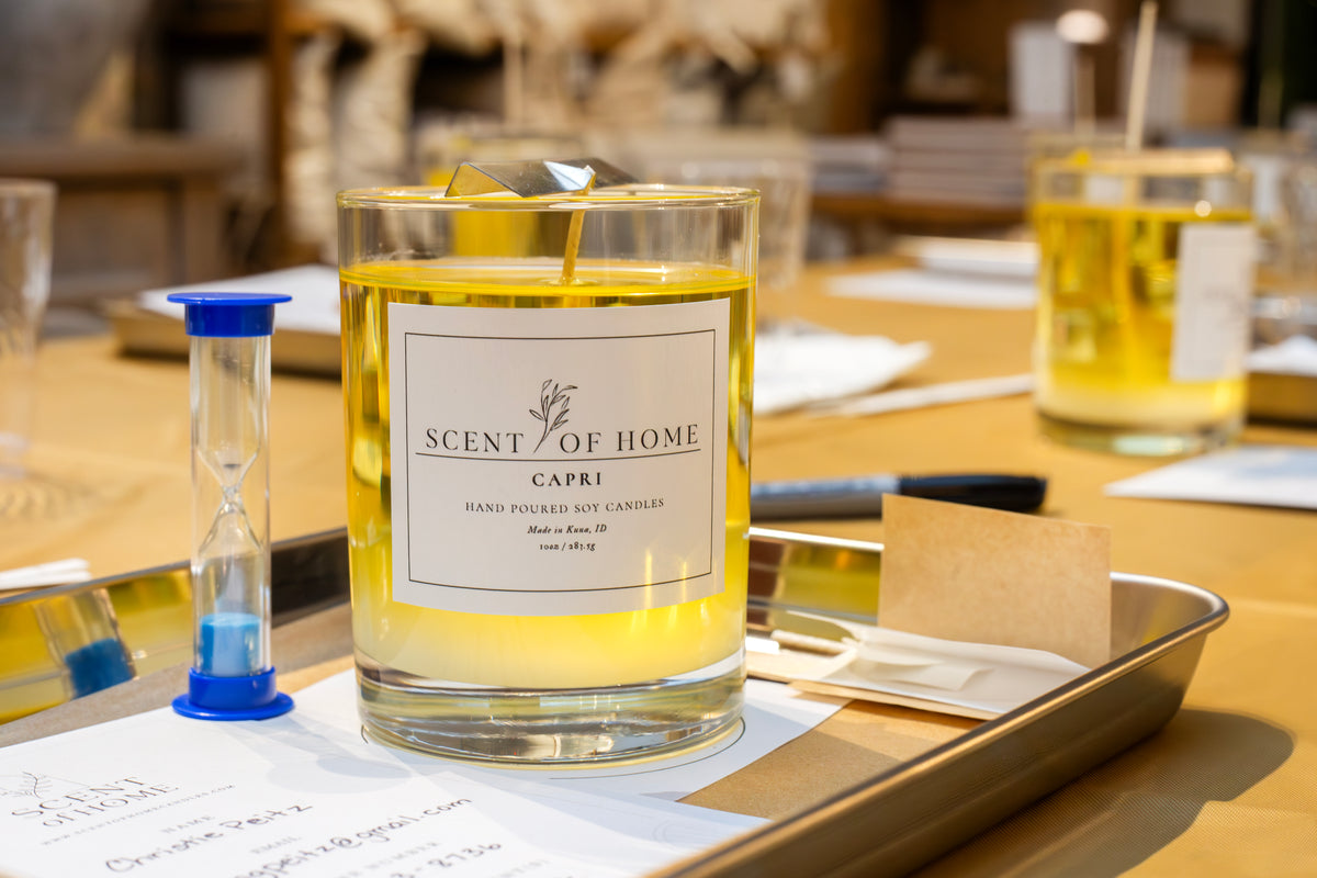 Close-up of a freshly poured Scent of Home soy candle labeled “Capri” cooling on a metal tray during a candle pouring class, surrounded by workshop tools, notes, and a small hourglass timer.