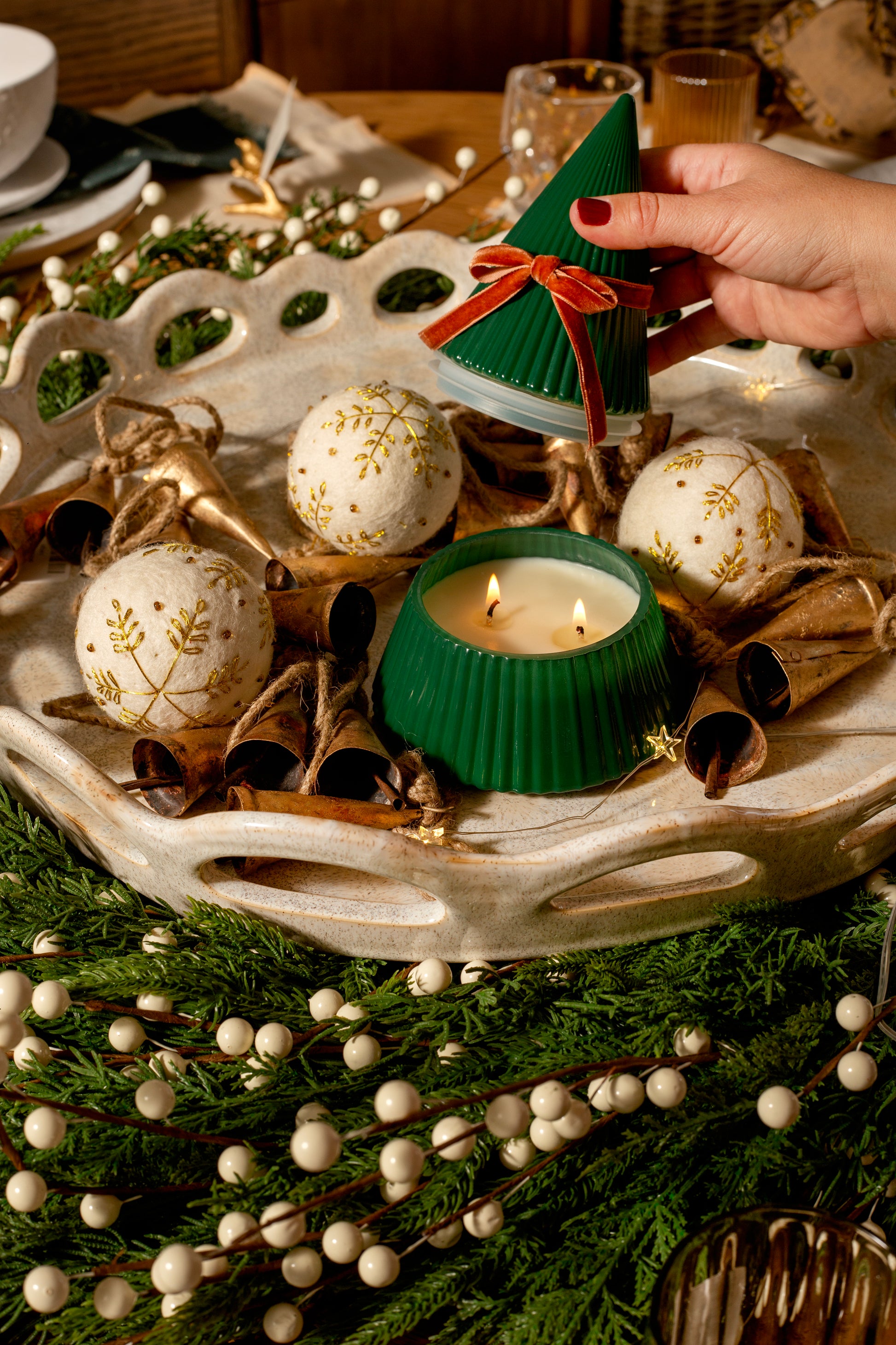 Hand lifting the lid of the Frosted Juniper Christmas Tree candle, revealing the lit candle inside, styled with holiday greenery, bells, and ornaments on a decorative tray.