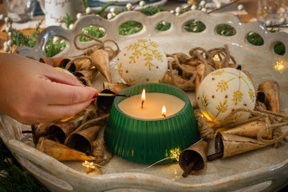 Frosted Juniper Christmas Tree candle burning in its green tree-shaped vessel, surrounded by holiday décor, while a hand lights the wick.