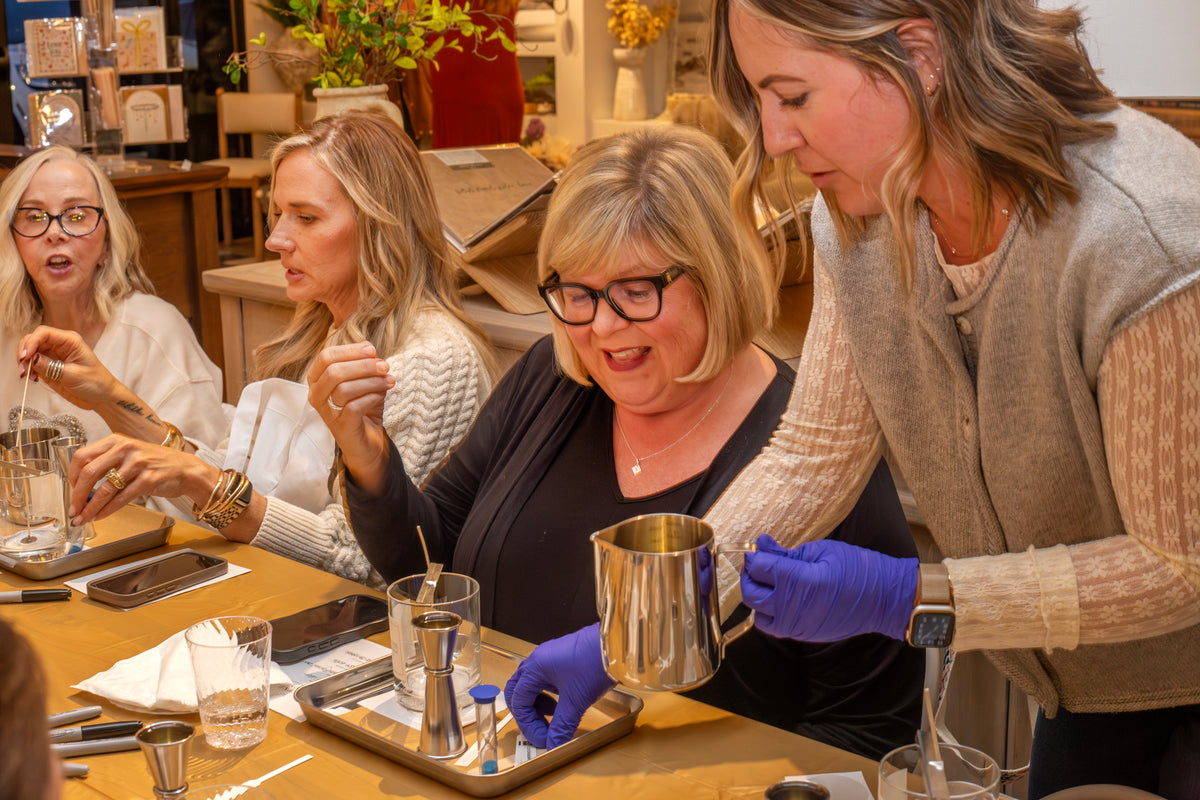 Jessica from Scent of Home assists participants during a candle pouring class, guiding them through the process of mixing and pouring wax at a warmly lit workshop table filled with supplies and glass jars.