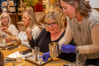 Jessica from Scent of Home assists participants during a candle pouring class, guiding them through the process of mixing and pouring wax at a warmly lit workshop table filled with supplies and glass jars.
