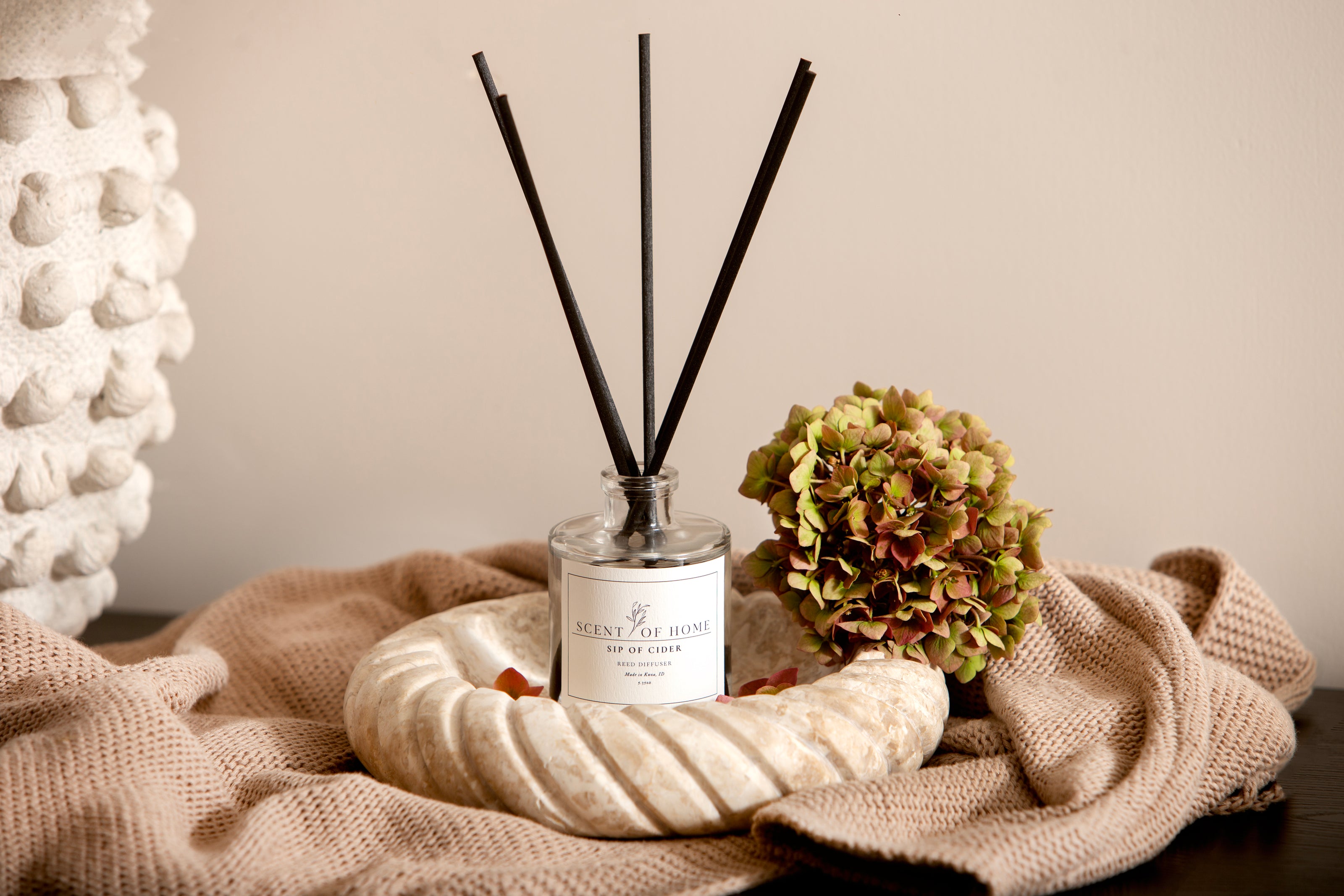 Scent of Home Sip of Cider reed diffuser displayed on a neutral knit blanket with hydrangea and ceramic décor — showcasing the warm, elegant style of the reed diffuser collection
