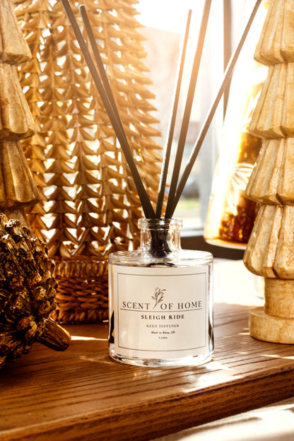 Scent of Home Sleigh Ride reed diffuser displayed on a wooden shelf, surrounded by warm gold holiday decor and sunlight.