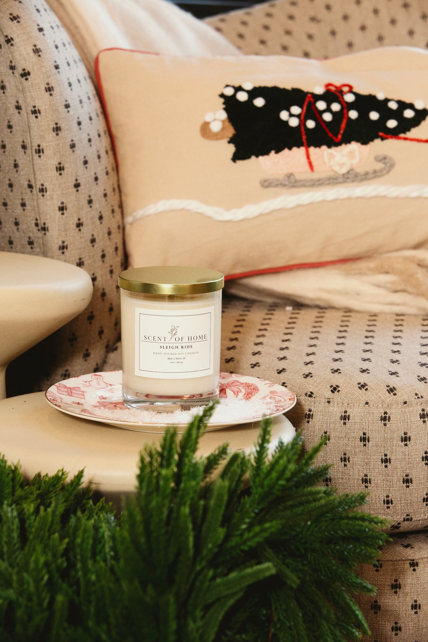 Scent of Home candle displayed on a decorative plate, set on a cozy upholstered chair with a holiday pillow and greenery in the foreground.