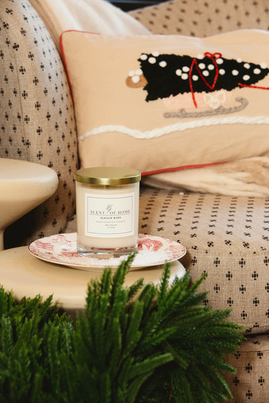 Scent of Home candle displayed on a decorative plate, set on a cozy upholstered chair with a holiday pillow and greenery in the foreground.