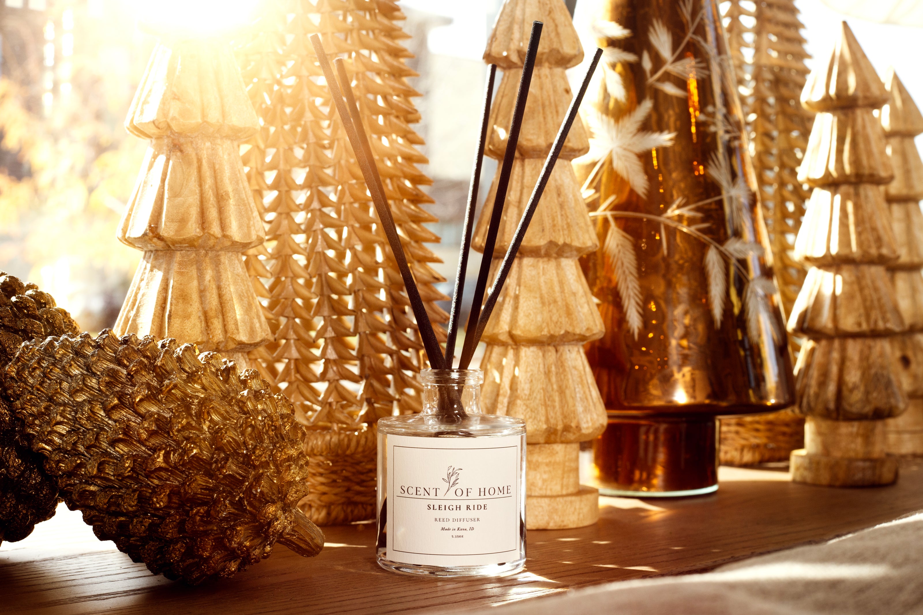 Scent of Home Sleigh Ride reed diffuser surrounded by gold-toned holiday trees and pinecones, bathed in warm sunlight for a cozy, festive glow.
