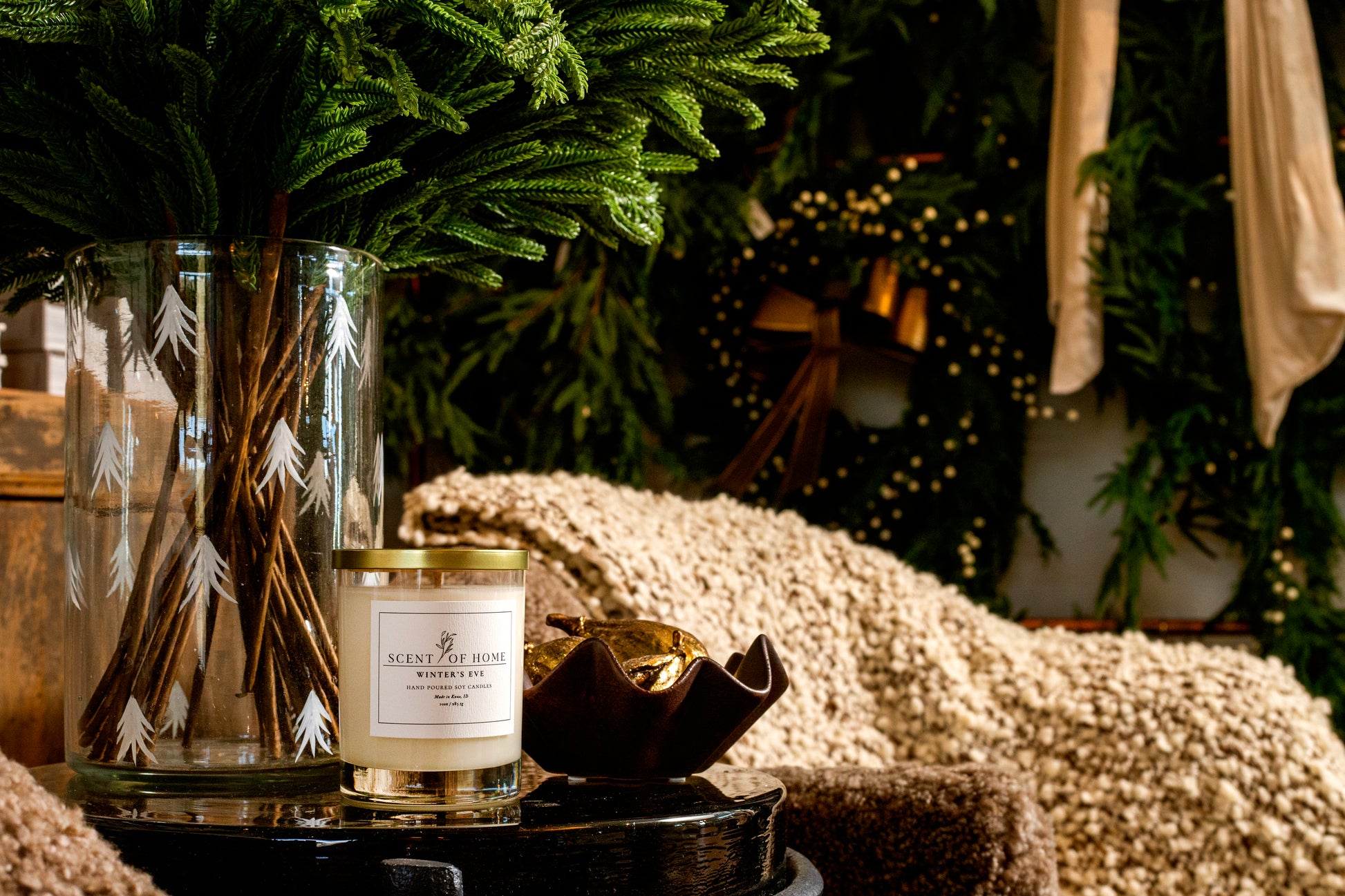 Scent of Home Winter’s Eve candle displayed beside a tall glass vase filled with evergreen branches, set in a cozy holiday scene with soft blankets and festive greenery in the background.