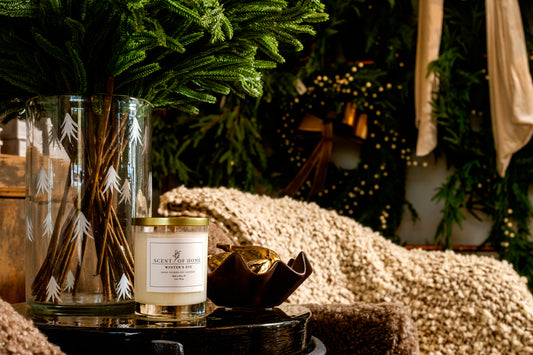 Scent of Home Winter’s Eve candle displayed beside a tall glass vase filled with evergreen branches, set in a cozy holiday scene with soft blankets and festive greenery in the background.