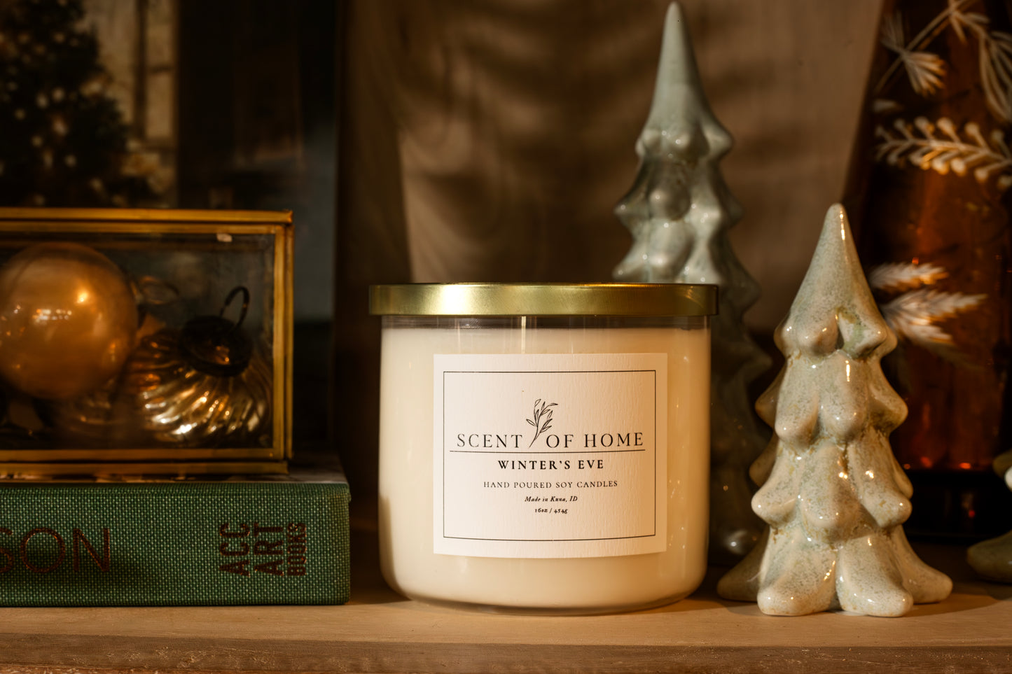 Scent of Home Winter’s Eve 3-wick soy candle displayed on a shelf with ceramic holiday trees, vintage ornaments, and a green book.