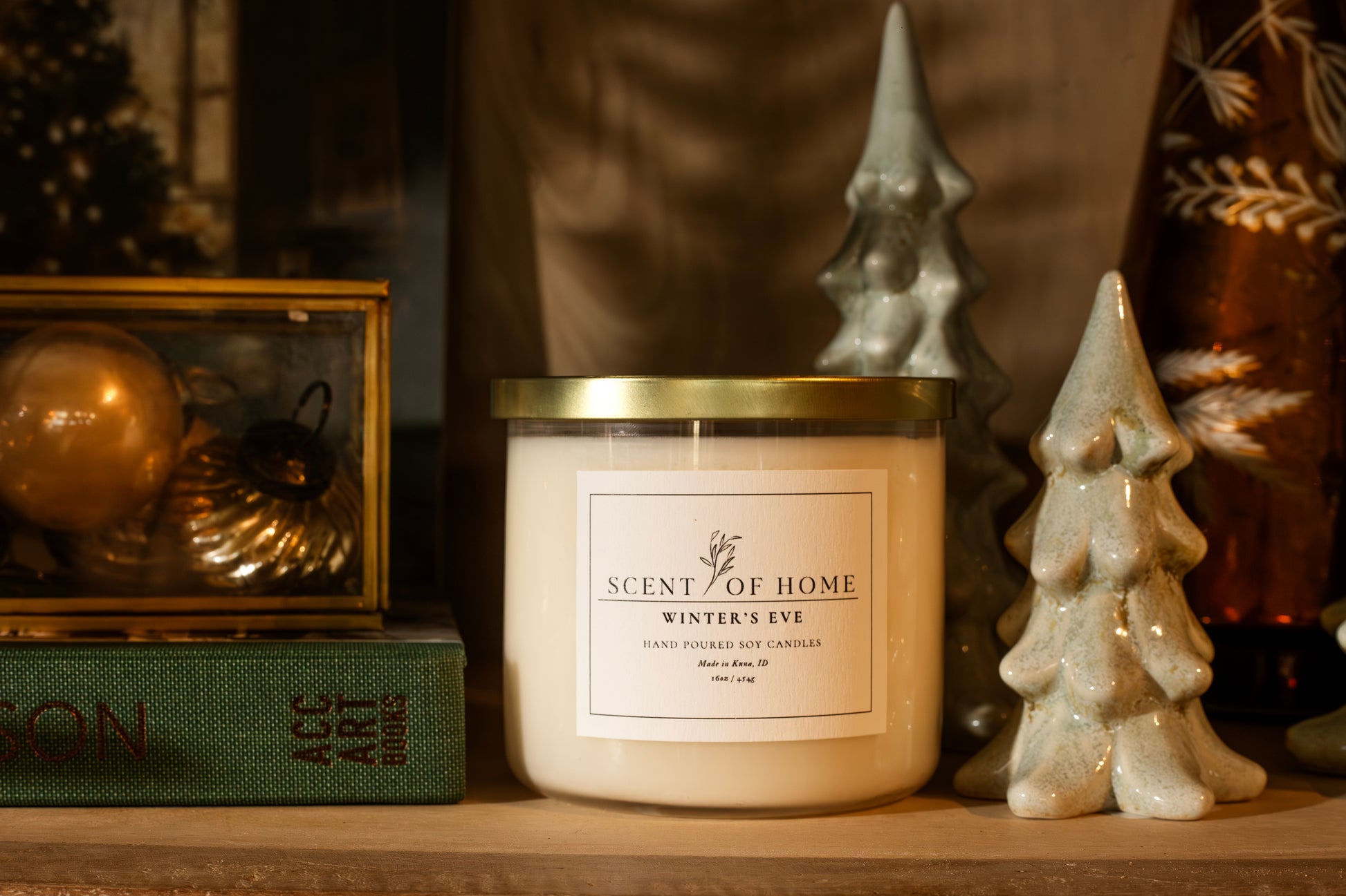 Scent of Home Winter’s Eve 3-wick soy candle displayed on a shelf with ceramic holiday trees, vintage ornaments, and a green book.