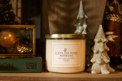 Scent of Home Winter’s Eve 3-wick soy candle displayed on a shelf with ceramic holiday trees, vintage ornaments, and a green book.