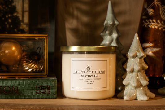 Scent of Home Winter’s Eve 3-wick soy candle displayed on a shelf with ceramic holiday trees, vintage ornaments, and a green book.