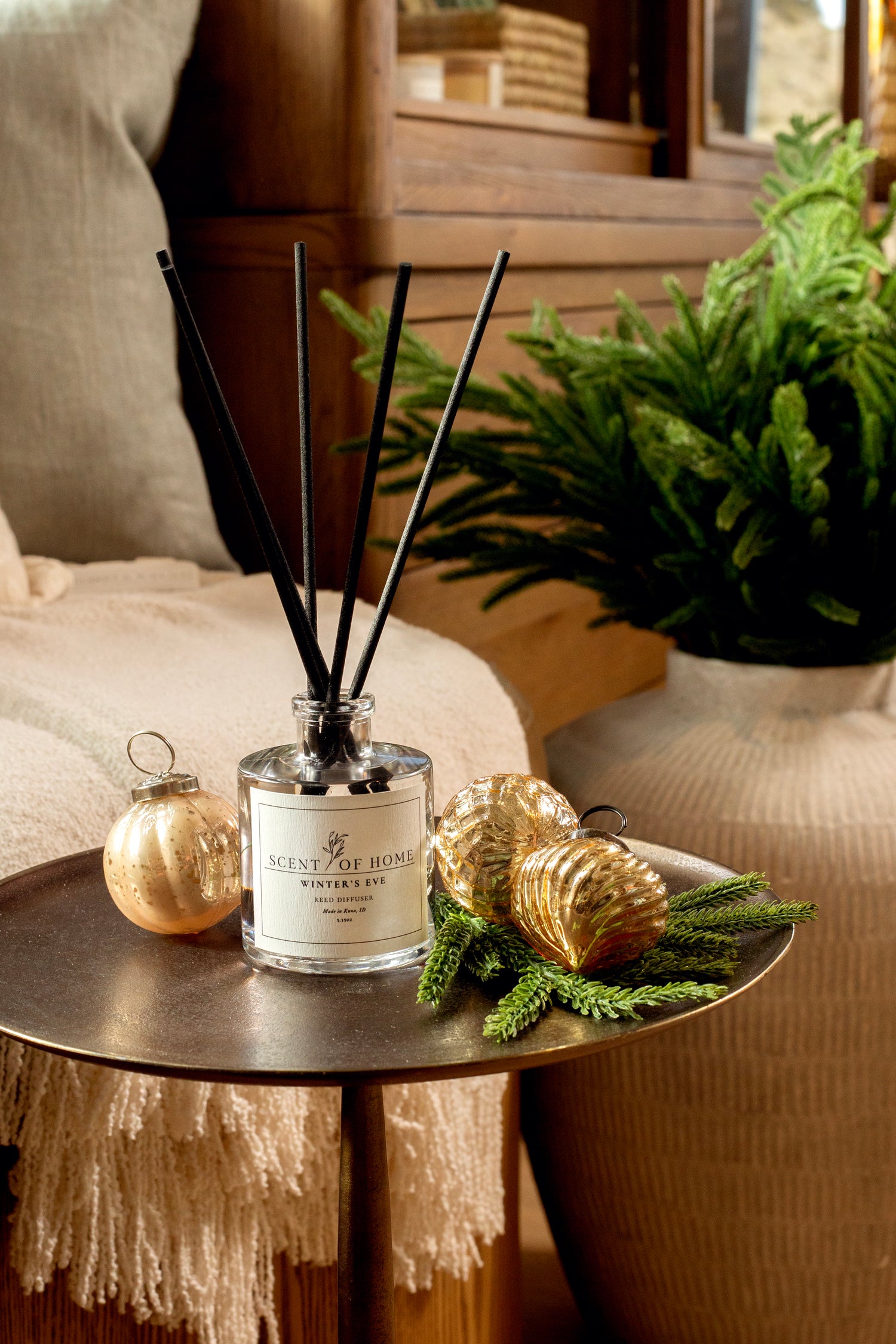 Scent of Home Winter’s Eve reed diffuser displayed on a side table with gold ornaments, evergreen sprigs, and a cozy throw blanket in a warm, inviting holiday setting.