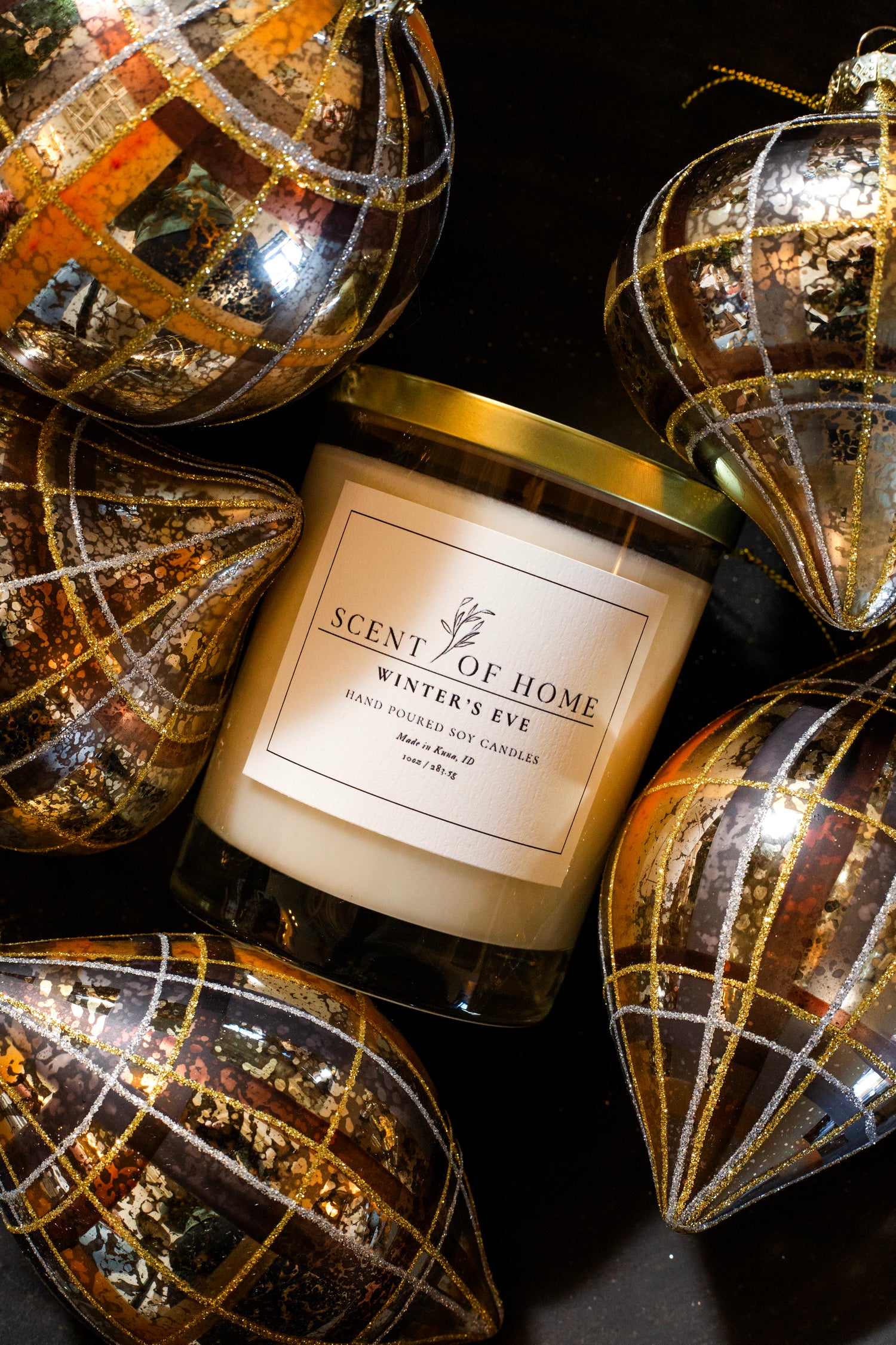 Scent of Home Winter’s Eve candle surrounded by gold and plaid-patterned glass ornaments, creating a rich and elegant holiday display.